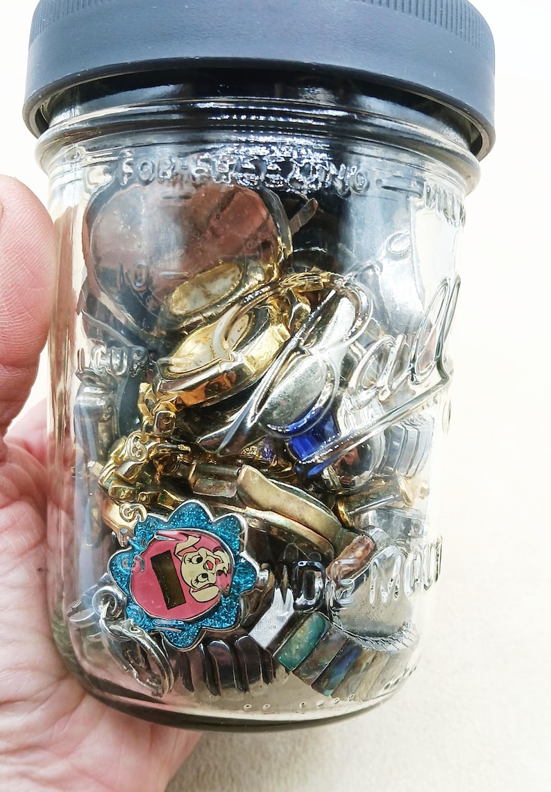 K&ouml;nnte beinhalten: Ein durchsichtiges Ball-Einmachglas aus Glas mit einem dunkelgrauen Deckel, gef&uuml;llt mit verschiedenen Metallgegenst&auml;nden, darunter goldfarbene Uhrenteile und ein blau glitzernder Anh&auml;nger mit einer rosa Zeichentrickfigur.