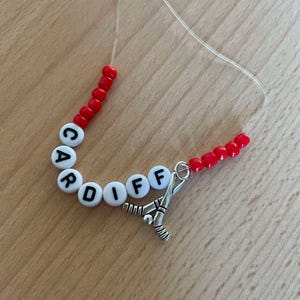 May include: A red and white beaded necklace with the word "CARDIFF" spelled out in white beads. A silver hockey stick charm is attached to the end of the necklace.