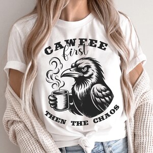 May include: White t-shirt with a black graphic of a raven holding a coffee mug. The text on the shirt reads "CAWFEE first THEN THE CHAOS". The raven is depicted with steam rising from the coffee.