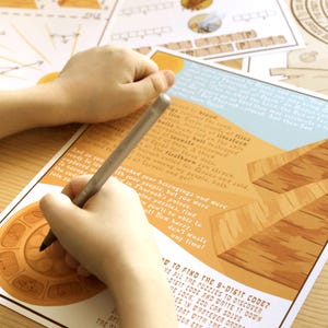 May include: A person's hands writing on a printed game board with a pen. The board features illustrations of pyramids and text. The game board is on a light-colored wooden surface. Additional game pieces are visible in the background.