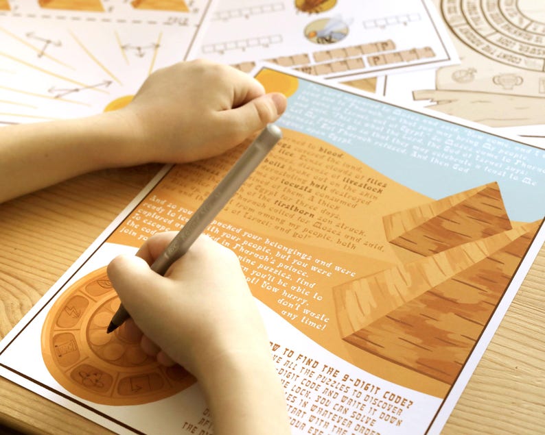 May include: A person's hands writing on a printed game board with a pen. The board features illustrations of pyramids and text. The game board is on a light-colored wooden surface. Additional game pieces are visible in the background.