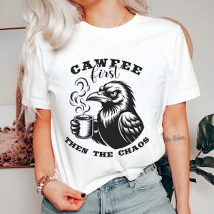 May include: White t-shirt with a black graphic of a raven holding a coffee mug. The text "CAWFEE first THEN THE CHAOS" is printed above and below the raven illustration. The shirt is a casual style.