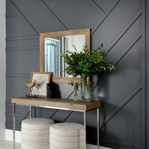 K&ouml;nnte beinhalten: Eine dunkelgraue Wand mit geometrischem Muster, ein Konsolentisch aus Holz mit einem Spiegel dar&uuml;ber und zwei wei&szlig;e Hocker. Der Konsolentisch hat eine Holzplatte und silberne Metallbeine. Der Spiegel hat einen Holzrahmen. Auf dem Tisch stehen Pflanzen in einer Vase.