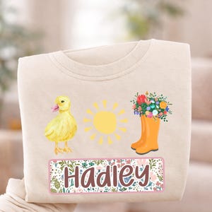 May include: Cream-colored sweatshirt featuring a yellow duckling, a yellow sun, and an orange rain boot filled with flowers. The name "Hadley" is displayed in a floral frame. A cheerful design for spring or summer.