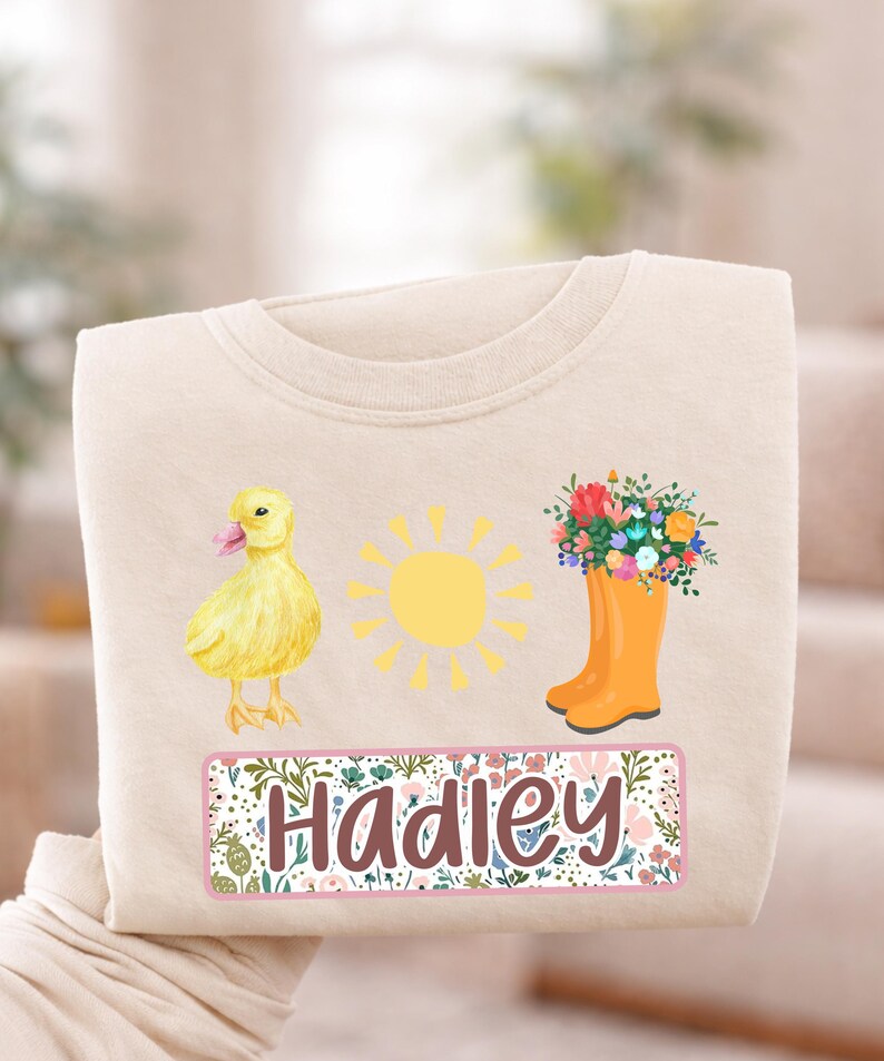 May include: Cream-colored sweatshirt featuring a yellow duckling, a yellow sun, and an orange rain boot filled with flowers. The name "Hadley" is displayed in a floral frame. A cheerful design for spring or summer.