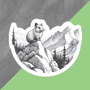 Puede incluir: Adhesivo en blanco y negro que representa una pika posada en una cima rocosa, con una cordillera y árboles de hoja perenne en el fondo. La pegatina tiene un borde blanco y está sobre un fondo gris y verde.