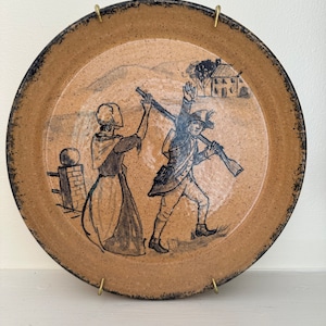 Peut inclure: Une assiette en céramique avec une scène peinte à la main d'une femme et d'un homme en vêtements coloniaux. L'homme tient un fusil et la femme tient un bâton. La scène se déroule dans un paysage rural avec une maison en arrière-plan.