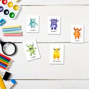 May include: Five cards with watercolor monster illustrations, each depicting a different emotion. The monsters are in various colors, including blue, purple, orange, and green. Text in German accompanies each monster, such as "Gl&uuml;cklich" and "Stolz". The cards are arranged on a white wooden surface with art supplies.