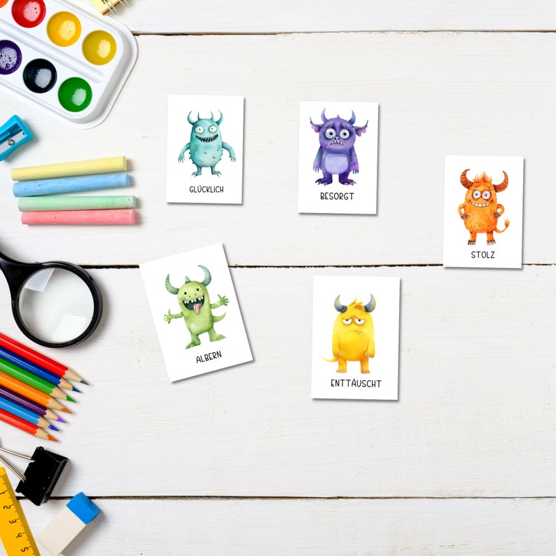 May include: Five cards with watercolor monster illustrations, each depicting a different emotion. The monsters are in various colors, including blue, purple, orange, and green. Text in German accompanies each monster, such as "Gl&uuml;cklich" and "Stolz". The cards are arranged on a white wooden surface with art supplies.