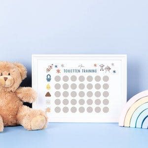 K&ouml;nnte beinhalten: Ein wei&szlig;er gerahmter Chart mit dem Text "Mein Toiletten Training" und einem Raster mit 36 Kreisen. Der Chart hat eine Rakete, eine Toilette, ein Kack-Emoji, ein H&auml;ndewaschen-Symbol und ein Raumschiff darauf. Ein Teddyb&auml;r und ein Regenbogenspielzeug befinden sich im Vordergrund.