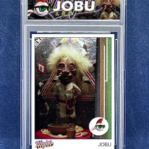 Puede incluir: Una tarjeta de béisbol de Jobu en un estuche protector. La tarjeta presenta una imagen detallada de una muñeca Jobu con un atuendo blanco, con un pequeño cuenco y una taza. La tarjeta tiene el texto "Major League" y "Jobu".