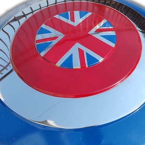 May include: A circular shield with a blue, silver, red, and white design. The centre features a red circle with a Union Jack flag. The shield has a reflective, metallic finish.