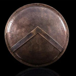May include: A bronze-colored, round shield with a V-shaped design. The shield has a metallic sheen and is likely made of metal. The shield has a dark background and is likely a historical or decorative item.