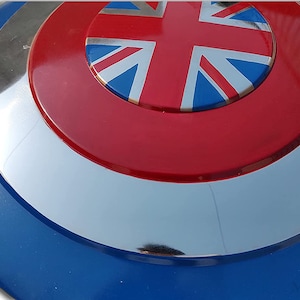 May include: A circular shield in blue, silver, and red. The centre displays a Union Jack on a red background. The shield has a reflective, metallic finish and a blue outer edge.