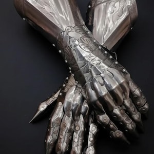 Può includere: Un paio di guanti ornati in metallo color argento, probabilmente per un'armatura. I guanti presentano intricati disegni floreali e a volute, con dita articolate che terminano con punte affilate. I rivetti fissano le piastre.