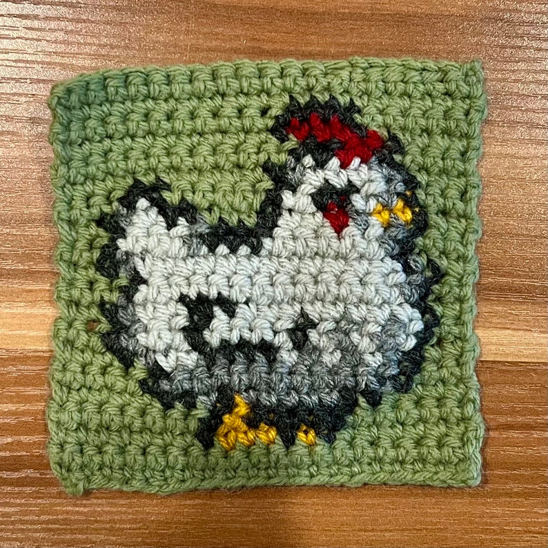 Puede incluir: Un cuadrado de crochet verde con un pollo de crochet blanco, gris, rojo y amarillo en el centro.