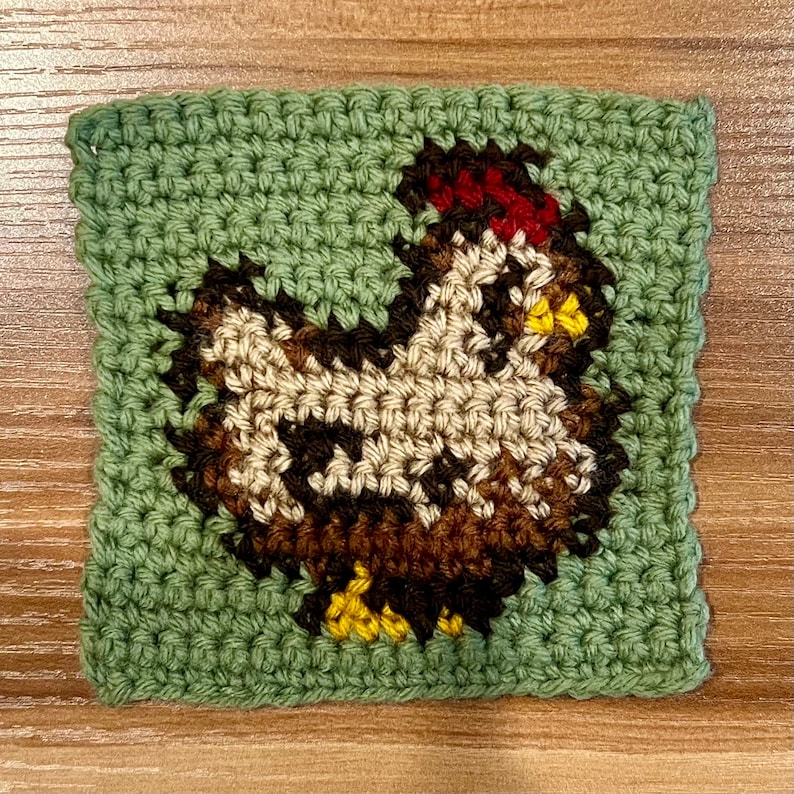 Puede incluir: Un cuadrado de crochet verde con un pollo de crochet marr&oacute;n, blanco y amarillo en el centro. El pollo tiene una cresta roja de crochet.