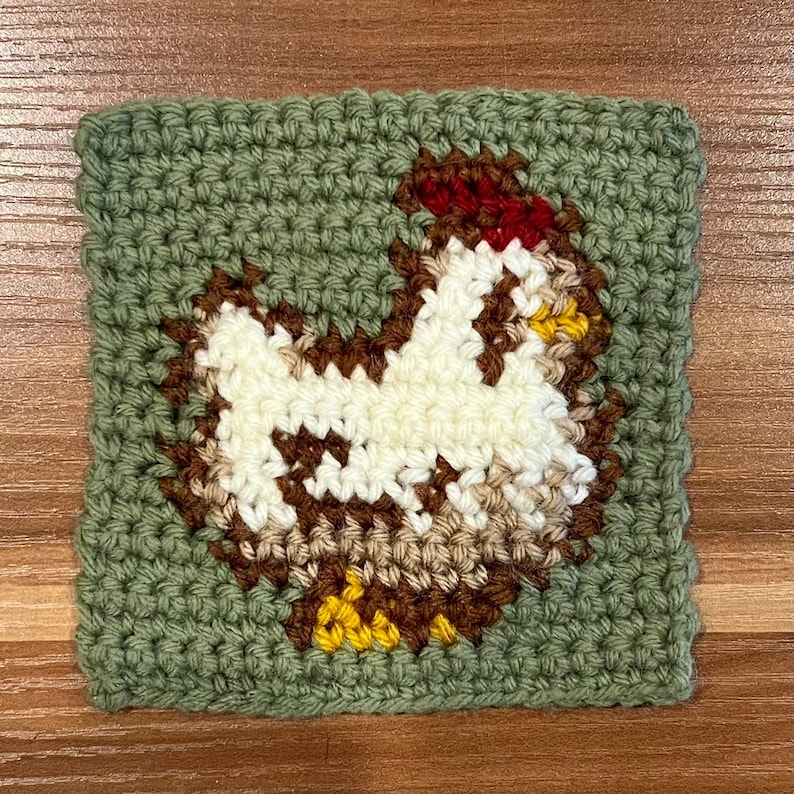 Puede incluir: Un cuadrado de crochet verde con un pollo de crochet blanco en el centro. El pollo tiene una cresta roja, plumas marrones y patas amarillas.