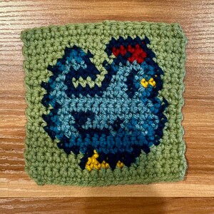 Puede incluir: Un cuadrado tejido a crochet con un pollo azul con cresta roja, pico amarillo y patas amarillas. El pollo est&aacute; rodeado por un borde verde.