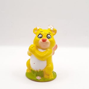 Op de afbeelding: Een geel Care Bear figuur met een roze vlinder op zijn rug. De beer heeft witte bloemen op zijn hoofd en staat op een groene basis met bloemen.