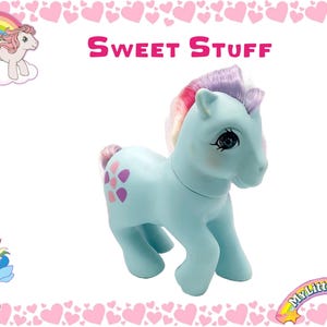 Puede incluir: Un juguete My Little Pony azul claro con una crin y cola rosa, morada y amarilla. El pony tiene un diseño de flor rosa en su flanco y las palabras "Sweet Stuff" están impresas sobre el pony.