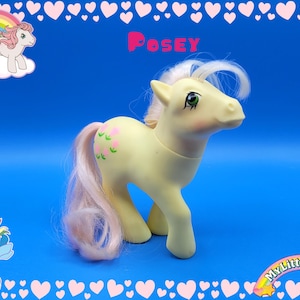 May include: A vintage My Little Pony toy named Posey, with a light yellow body, pink tail and mane, and green eyes. The pony has pink flower decals on its flank. The background is blue with heart and pony graphics.