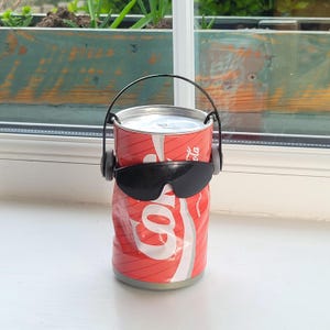 Puede incluir: Una lata de Coca-Cola reciclada con asa y gafas de sol negras. La lata roja y blanca tiene un aspecto aplastado, con el logotipo de Coca-Cola visible. Las gafas de sol están colocadas en el centro de la lata, dándole un aspecto divertido.