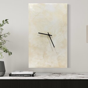 May include: A beige abstract wall clock with black hands, a black lamp with a beige shade, and two gray and black planters with green plants on a white marble countertop.
