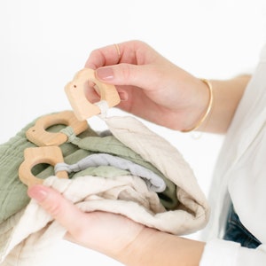 May include: A set of three wooden teethers attached to a stack of soft, green and beige muslin cloths. The teethers are shaped like a bird and have a ring for a baby to hold.