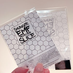 May include: A black and white sticker with the text "Shungite EMF Protection Slice" on a white background with a honeycomb pattern. The sticker is in a clear plastic bag with a black adhesive backing.
