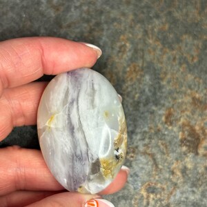 Puede incluir: Una piedra pulida lisa de forma ovalada con un patrón de mármol blanco, morado y marrón.