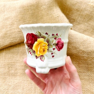 May include: A white ceramic planter featuring a floral pattern with a large central yellow rose, a deep red rose to the left, and a smaller pink rose to the right, accented by green leaves and small red bell-shaped flowers on a fluted rim base.