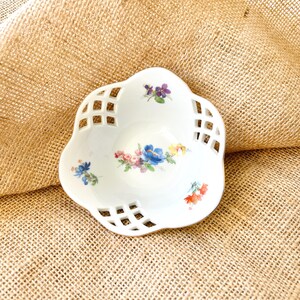 May include: A white, decorative, ceramic dish with a floral design. The dish has a scalloped edge with a lattice pattern and is adorned with colorful flowers in shades of blue, pink, and purple. The dish is approximately 6 inches in diameter.