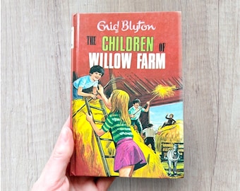 May include: A vintage hardcover book titled "The Children of Willow Farm" by Enid Blyton. The cover features a colorful illustration of children playing on a farm, with a hay bale and a white horse. The book is held by a hand.