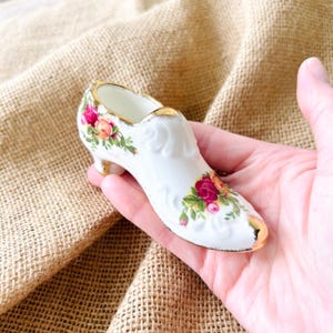 May include: A small white porcelain shoe figurine featuring embossed scrollwork details, a gold-painted rim, and colorful floral patterns of red and yellow roses on the toe and heel sections, finished with a gold-trimmed small heel.