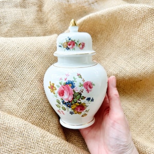 May include: White porcelain jar with a lid and floral design. The jar has a gold trim and features pink, yellow, and blue flowers.
