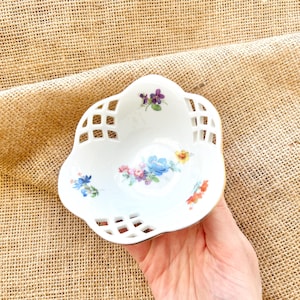 May include: White porcelain dish with a lattice design and floral accents in shades of blue, purple, pink, and yellow. The dish has a scalloped edge and is held by a hand.