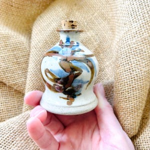 May include: A small, hand-painted ceramic bottle with a cork stopper. The bottle has a white base with abstract blue, brown, and tan brushstrokes. The cork is light brown. The bottle is held in a hand against a burlap background.