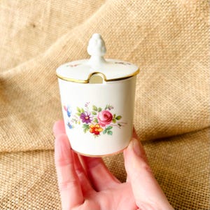 May include: A white ceramic sugar bowl with a lid and a gold rim. The bowl is decorated with a floral pattern of pink, purple, and yellow flowers. The lid has a small handle and a slot for a spoon.