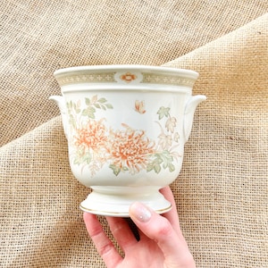 May include: A white ceramic planter with a floral design featuring orange and green flowers. The planter has two handles and a pedestal base.
