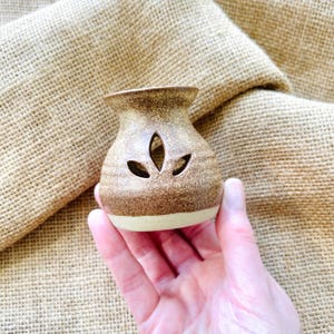 May include: A small, bulbous ceramic oil burner with a speckled brown and tan glaze. The vessel features a wide, flared rim and a cutout design on the front consisting of three leaf-shaped apertures arranged in a cluster to allow light to pass.