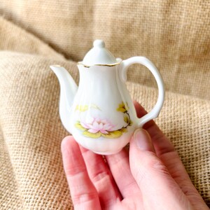 May include: A small, white ceramic teapot with a delicate floral design featuring a pink lotus flower and yellow accents. The teapot has a gold trim and a curved handle. The teapot is held in a hand.