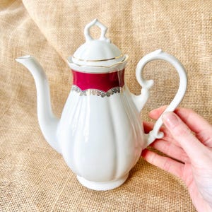 May include: White porcelain coffee pot with a red band and gold trim. The handle is curved, and the lid has a decorative finial. The pot has a ribbed design and a long spout. A hand is holding the pot.