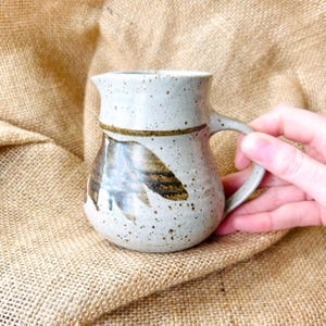 May include: A speckled, off-white ceramic jug with a leaf design in brown and tan. The jug has a handle and a spout. The jug is approximately 13 cm tall.