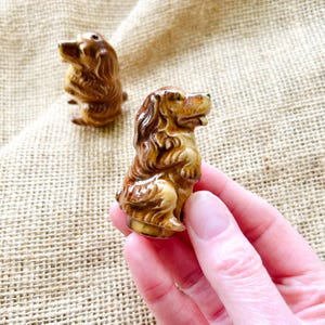 May include: Two small, brown ceramic dog figurines. The dogs are in a sitting position with detailed fur and facial features. One is in focus, held by a hand, while the other is slightly out of focus in the background. The figurines are on a textured, tan surface.