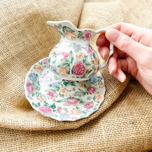 May include: A white ceramic creamer with a floral pattern in shades of pink, yellow, and blue. The creamer has a matching saucer with a scalloped edge.