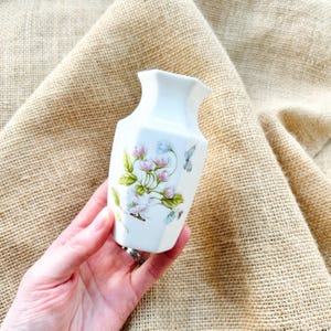 May include: A small white porcelain vase with a floral design featuring pink and white flowers and green leaves. A blue butterfly is also depicted on the vase.