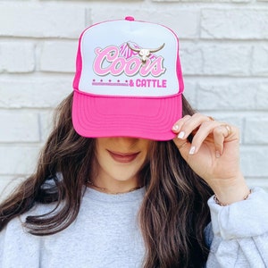May include: Pink trucker hat with a white mesh back. The hat has a white logo with the text "Coors & Cattle" and a stylized bull's head.