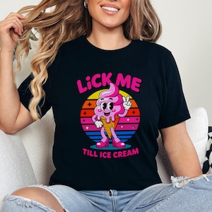 May include: Black t-shirt with a colorful graphic of a smiling ice cream cone giving a peace sign. The text "LICK ME" is above the ice cream cone, and "TILL ICE CREAM" is below. The ice cream cone is pink and wearing pink shoes.