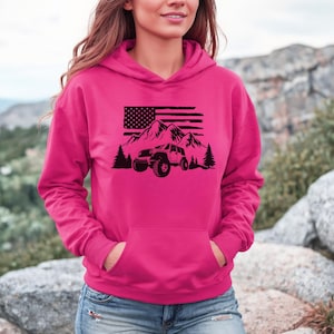 Puede incluir: Una sudadera con capucha rosa con un gráfico negro de un jeep conduciendo por un paisaje de montaña con una bandera estadounidense en el fondo.